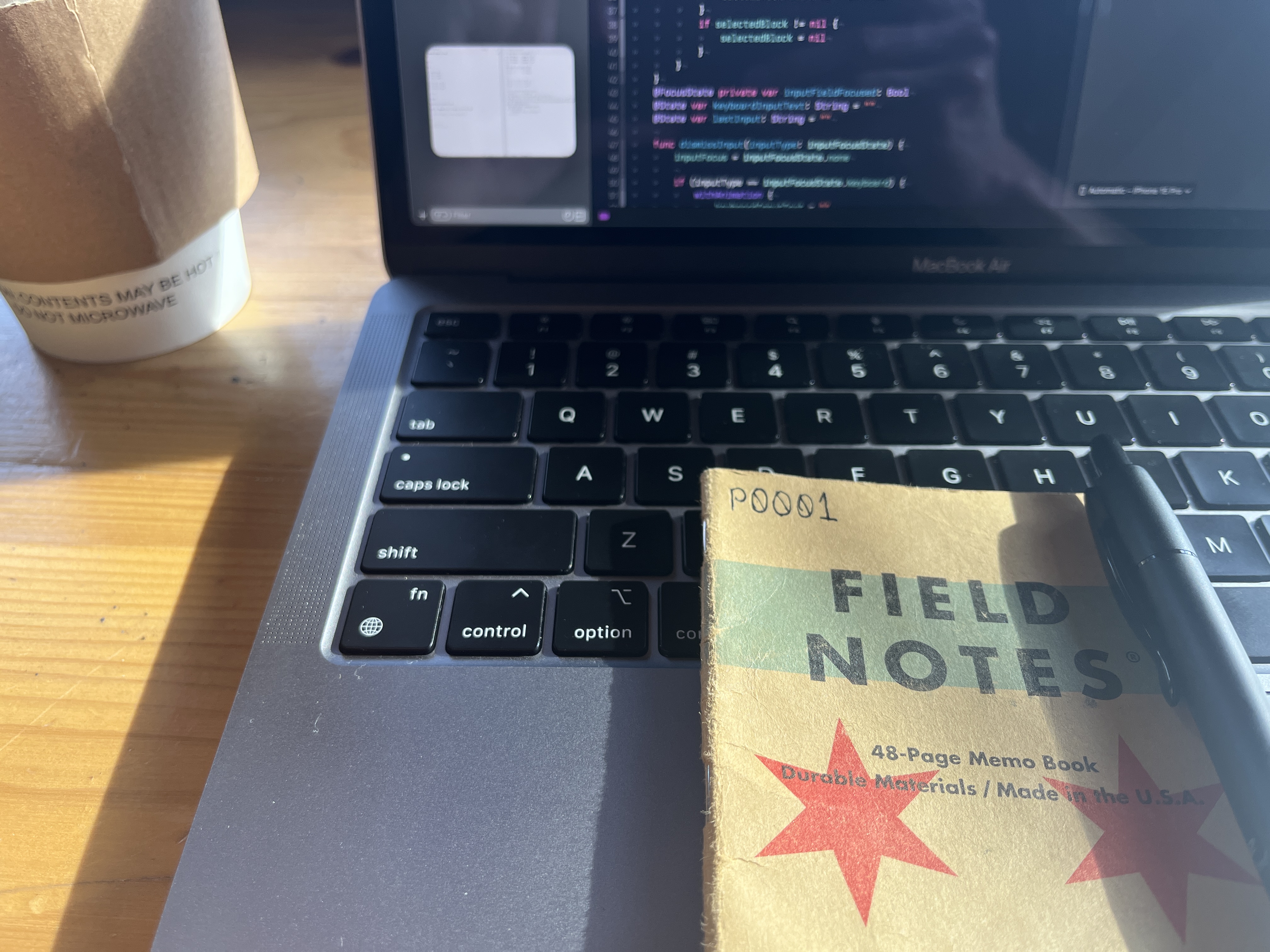 A work environment with a MacBook Air displaying code on the screen, a coffee cup labeled “CONTENTS MAY BE HOT”, and a “FIELD NOTES” memo book with a pen on a wooden table.